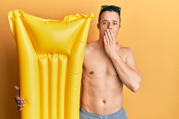 Handsome young man wearing swimsuit and holding summer float covering mouth with hand, shocked and afraid for mistake. surprised expression  - Photo, Image