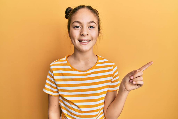 Красивая брюнетка маленькая девочка в повседневной полосатой футболке с большой улыбкой на лице, указывая пальцем руки в сторону, глядя в камеру.  - Фото, изображение