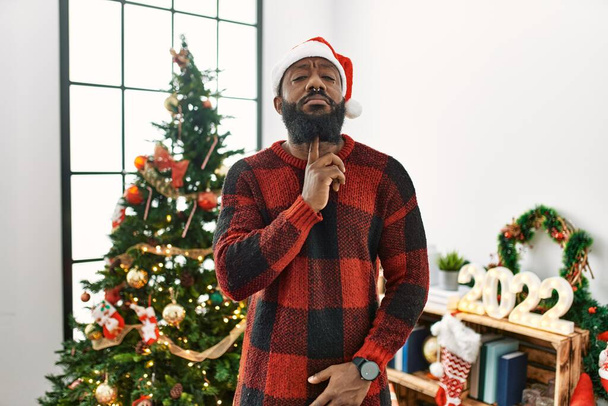 Αφροαμερικάνος που φοράει καπέλο Σάντα Κλάους στέκεται δίπλα στο χριστουγεννιάτικο δέντρο σκεπτόμενος συγκεντρωμένος για την αμφιβολία με το δάχτυλο στο πηγούνι και κοιτάζοντας προς τα πάνω αναρωτιέται  - Φωτογραφία, εικόνα