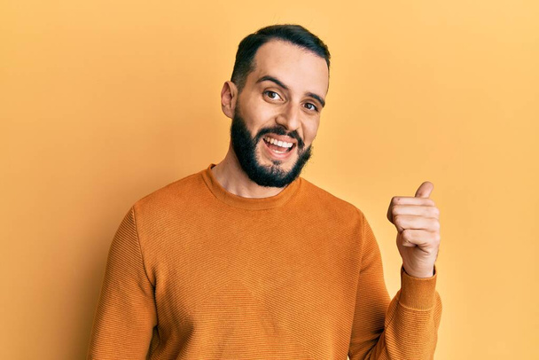 Young man with beard wearing casual winter sweater smiling with happy face looking and pointing to the side with thumb up.  - Foto, immagini