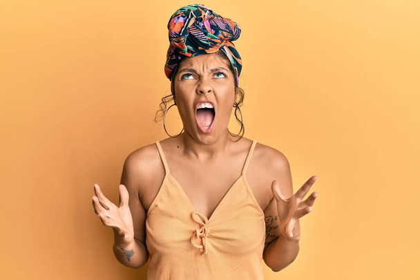 Young hispanic girl wearing hair turban over yellow background crazy and mad shouting and yelling with aggressive expression and arms raised. frustration concept.  - Фото, изображение
