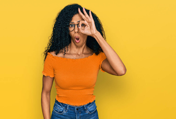 Middle age african american woman wearing casual clothes and glasses doing ok gesture shocked with surprised face, eye looking through fingers. unbelieving expression.  - Photo, Image