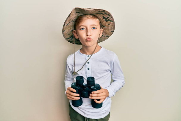 Petit garçon caucasien portant un chapeau explorateur tenant des jumelles regardant la caméra souffler un baiser étant charmant et sexy. expression d'amour.  - Photo, image