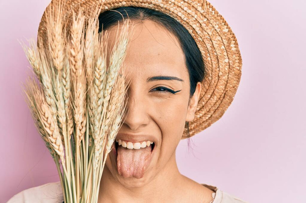 Joven chica hispana con sombrero de verano sosteniendo espiga de trigo sobre el ojo sacando la lengua feliz con expresión divertida.  - Foto, Imagen