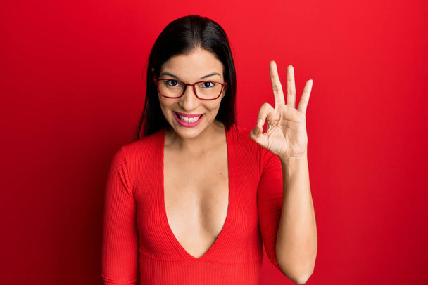 Junge lateinamerikanische Frau in lässiger Kleidung und Brille, die positiv lächelt und mit Händen und Fingern Okay-Zeichen macht. Erfolgreicher Ausdruck.  - Foto, Bild