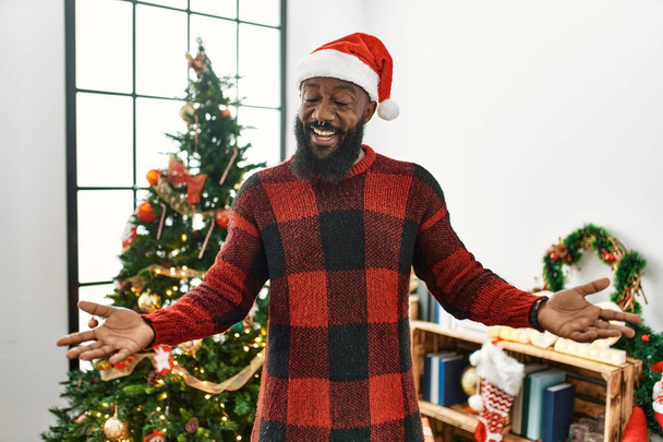 Αφροαμερικάνος άνδρας φορώντας καπέλο santa claus στέκεται από το χριστουγεννιάτικο δέντρο χαμογελώντας χαρούμενα με ανοιχτές αγκάλες ως φιλικό καλωσόρισμα, θετική και αυτοπεποίθηση χαιρετισμούς  - Φωτογραφία, εικόνα