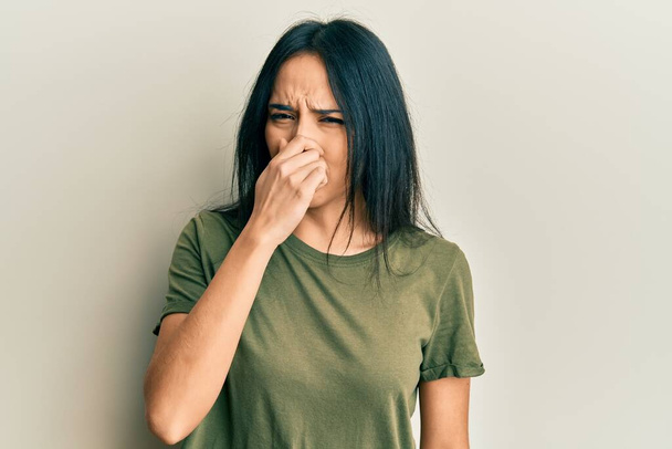 Young hispanic girl wearing casual t shirt smelling something stinky and disgusting, intolerable smell, holding breath with fingers on nose. bad smell  - Фото, изображение
