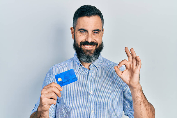 Genç İspanyol adam kredi kartı tutuyor. Parmaklarıyla imza atıyor, gülümsüyor. Mükemmel bir sembol.  - Fotoğraf, Görsel