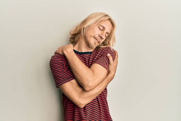 Καυκάσιος νεαρός άνδρας με μακριά μαλλιά που φορούσε casual ριγέ μπλουζάκι αγκαλιάζοντας τον εαυτό του χαρούμενο και θετικό, χαμογελώντας με αυτοπεποίθηση. αυτοαγάπη και αυτοφροντίδα  - Φωτογραφία, εικόνα