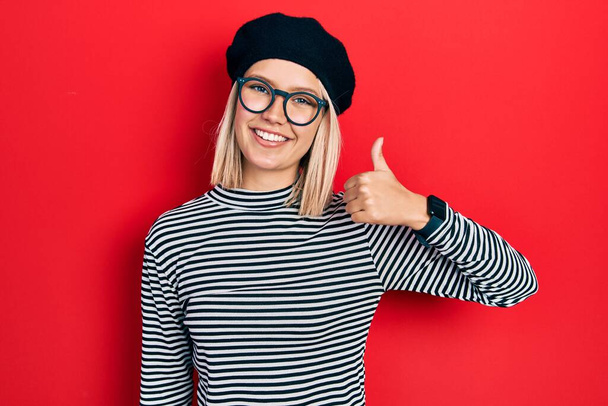 Piękna blondynka nosi francuski wygląd z beret okulary uśmiechnięty szczęśliwy i pozytywny, kciuk do góry robi doskonałe i znak aprobaty  - Zdjęcie, obraz