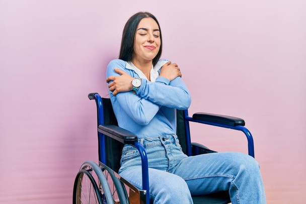 Женщина с голубыми глазами, сидящая на инвалидной коляске, обнимает себя счастливой и позитивной, улыбается уверенно. любовь к себе и забота о себе  - Фото, изображение