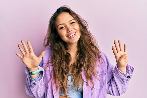 Junges hispanisches Mädchen trägt lässige Kleidung und zeigt mit Finger Nummer neun nach oben, während sie selbstbewusst und glücklich lächelt.  - Foto, Bild