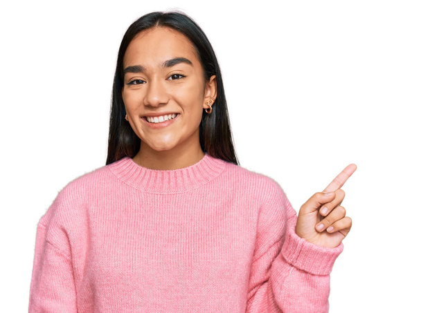 Joven mujer asiática vistiendo suéter casual de invierno con una gran sonrisa en la cara, señalando con el dedo de la mano a un lado mirando a la cámara.  - Foto, imagen