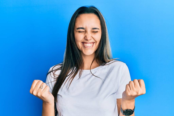 Νεαρή Ισπανίδα γυναίκα φορώντας casual λευκό t πουκάμισο πολύ χαρούμενος και ενθουσιασμένος κάνει χειρονομία νικητής με τα χέρια ψηλά, χαμογελώντας και ουρλιάζοντας για την επιτυχία. έννοια εορτασμού.  - Φωτογραφία, εικόνα