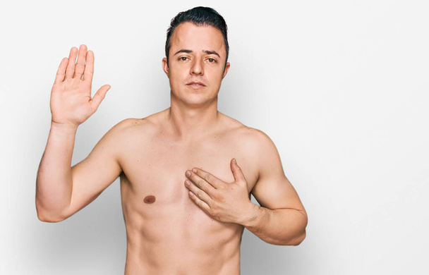 Bello giovane uomo che indossa costumi da bagno senza maglietta giurando con mano sul petto e palmo aperto, facendo un giuramento di fedeltà  - Foto, immagini