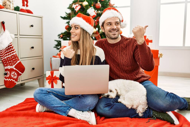Young couple of wife and husband wearing christmas hat using laptop pointing thumb up to the side smiling happy with open mouth  - Photo, Image