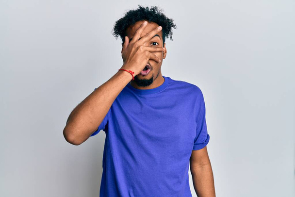 Young african american man with beard wearing casual blue t shirt peeking in shock covering face and eyes with hand, looking through fingers afraid  - Foto, afbeelding