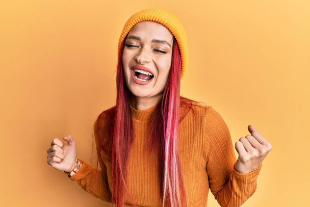 Young caucasian woman wearing wool cap very happy and excited doing winner gesture with arms raised, smiling and screaming for success. celebration concept.  - Foto, Imagen