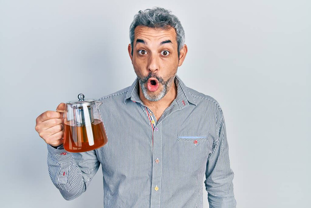 Handsome middle age man with grey hair holding traditional tea pot scared and amazed with open mouth for surprise, disbelief face  - Foto, immagini