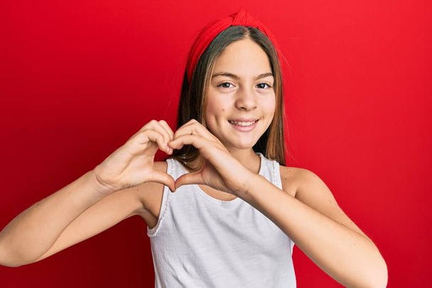 Beautiful brunette little girl wearing casual white t shirt smiling in love doing heart symbol shape with hands. romantic concept.  - Foto, afbeelding