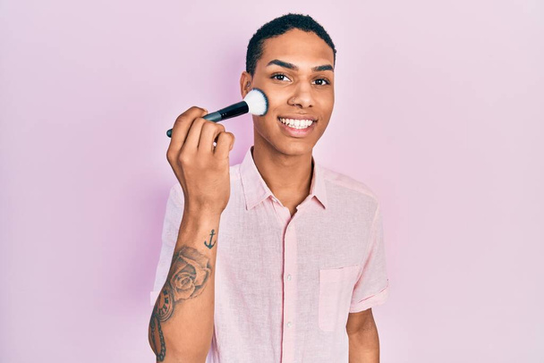 Young african american guy holding makeup brush looking positive and happy standing and smiling with a confident smile showing teeth  - Valokuva, kuva
