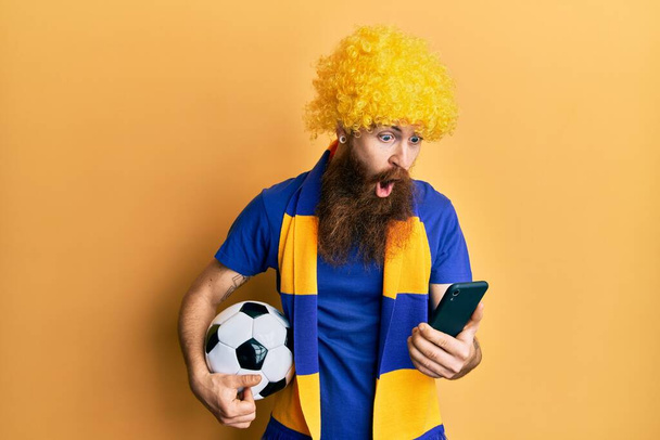 Redhead άνθρωπος με μακριά γενειάδα ποδόσφαιρο χούλιγκαν επευφημίες παιχνίδι κρατώντας smartphone φοβάται και σοκαρισμένος με έκπληξη και κατάπληκτος έκφραση, φόβο και ενθουσιασμένος πρόσωπο.  - Φωτογραφία, εικόνα