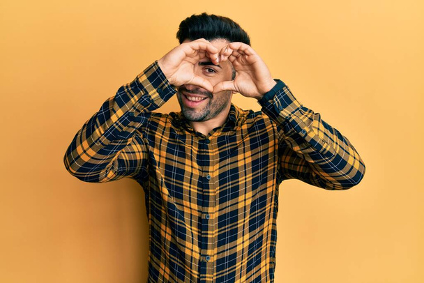 Joven hombre hispano usando ropa casual haciendo forma de corazón con la mano y los dedos sonriendo mirando a través del signo  - Foto, imagen