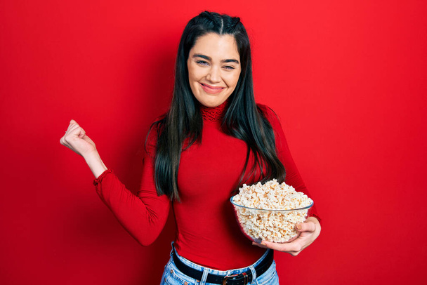 Молодая латиноамериканка ест попкорн крича гордо, празднуя победу и успех очень взволнован с поднятой рукой  - Фото, изображение