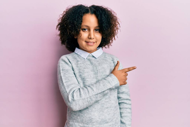 Маленькая девочка с афро-волосами в повседневной одежде улыбается весело указывая рукой и пальцем в сторону  - Фото, изображение