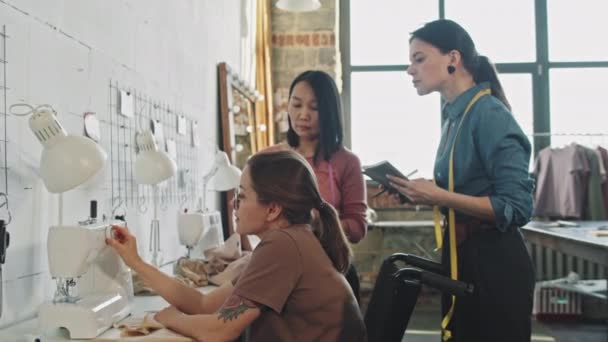 Tracking met slowmo van vrouwelijke naaister met behulp van naaimachine en praten met vrouwelijke modeontwerpers het maken van notities in de studio - Video