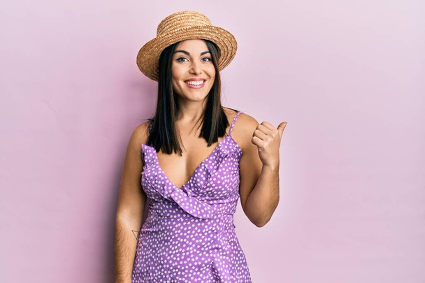 Junge brünette Frau in Sommerkleid und Hut lächelt mit glücklichem Gesicht und zeigt mit erhobenem Daumen zur Seite.  - Foto, Bild