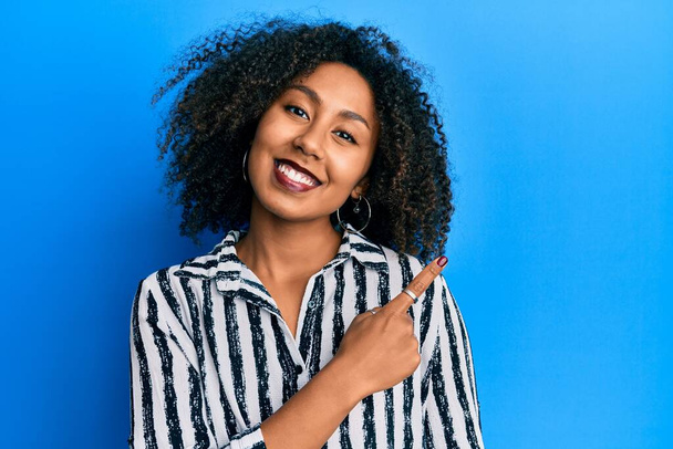 Belle femme afro-américaine avec des cheveux afro portant des vêtements décontractés souriant joyeuse pointant avec la main et le doigt sur le côté  - Photo, image