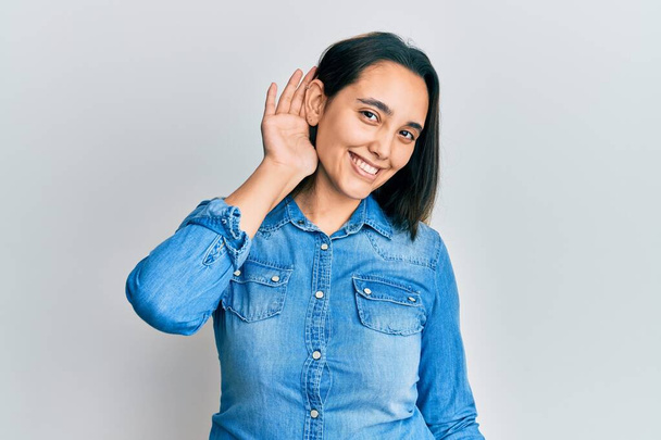 Νεαρή Ισπανίδα γυναίκα που φοράει απλό τζιν μπουφάν χαμογελώντας με το χέρι πάνω από το αυτί ακούγοντας μια ακρόαση για φήμες ή κουτσομπολιά. έννοια της κώφωσης.  - Φωτογραφία, εικόνα
