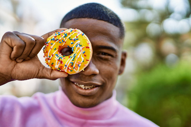 Genç Afrikalı Amerikalı adam gülümsüyor. Gözünün önünde donut tutarak şehre gülümsüyor.. - Fotoğraf, Görsel