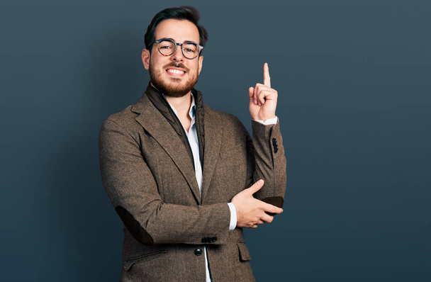 Νεαρός Ισπανός άνδρας με επιχειρηματικό σακάκι και γυαλιά με ένα μεγάλο χαμόγελο στο πρόσωπο, δείχνοντας με το χέρι και το δάχτυλο στο πλάι κοιτάζοντας την κάμερα.  - Φωτογραφία, εικόνα
