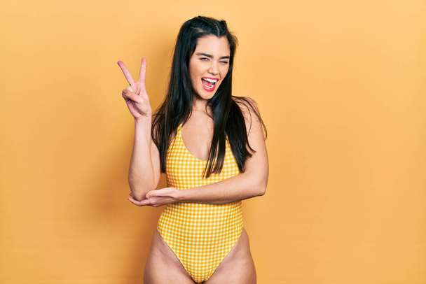 Young hispanic girl wearing swimsuit smiling with happy face winking at the camera doing victory sign with fingers. number two.  - Photo, Image