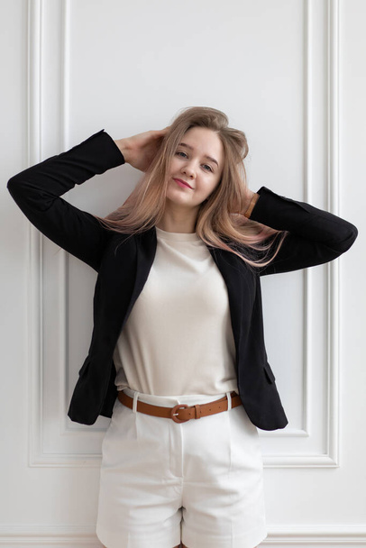 Beautiful young Caucasian woman in casual office clothes standing and touching her hair, posing, looking at camera. Vertical shot against white wall. - Photo, Image