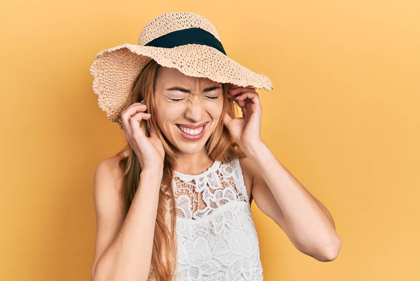Young caucasian woman wearing summer hat covering ears with fingers with annoyed expression for the noise of loud music. deaf concept.  - 写真・画像