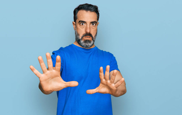 Muž středního věku s vousy v neformální modré tričko strach a strach s výrazem strachu zastavit gesto s rukama, křičí v šoku. koncepce paniky.  - Fotografie, Obrázek