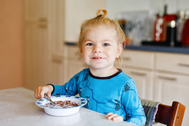 Küçük, güzel bir kız kahvaltıda sağlıklı mısır gevreği ve süt yiyor. Renkli giysiler içinde şirin bir bebek mutfakta oturup yulaf ve mısır gevreği hazırlarken eğleniyor. Evde kapalı. - Fotoğraf, Görsel