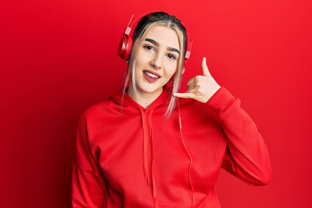 Молода сучасна дівчина носить спортивний одяг і використовує навушники, посміхаючись робити жест телефону з рукою і пальцями, як розмовляти по телефону. концепції спілкування
.  - Фото, зображення