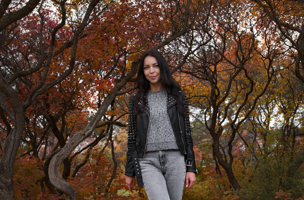 Jesienna młoda biała kobieta w czarnej skórzanej kurtce, szarym swetrze i dżinsach w jesiennym parku. Ciepła pogoda. - Zdjęcie, obraz