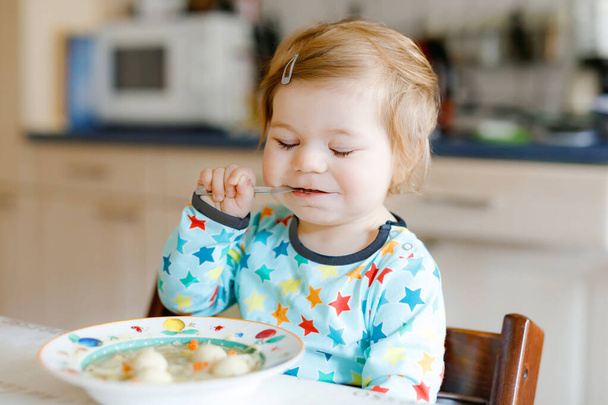 Очаровательная малышка ест с ложки овощного супа с лапшой. питание, ребенок, питание и концепция развития. Симпатичный малыш, дочь с ложкой сидит в кресле и учится есть самостоятельно. - Фото, изображение