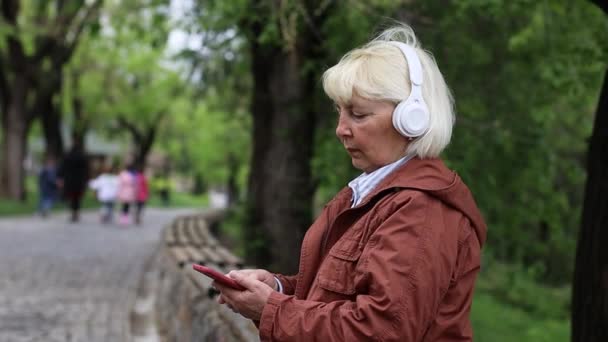 Retrato de anciana adulta de 50 años usando smartphone y escuchando música con auriculares en la calle al aire libre - Metraje, vídeo