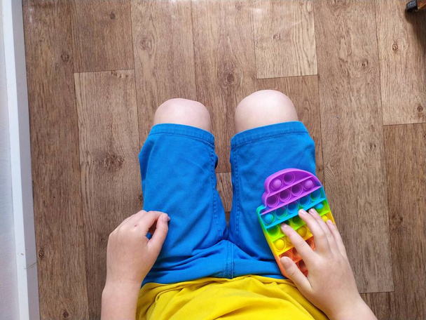 Boy in blue shorts play with colorful poppit fidget toy. Anti-stress sensory toy for kids and adults - Photo, Image