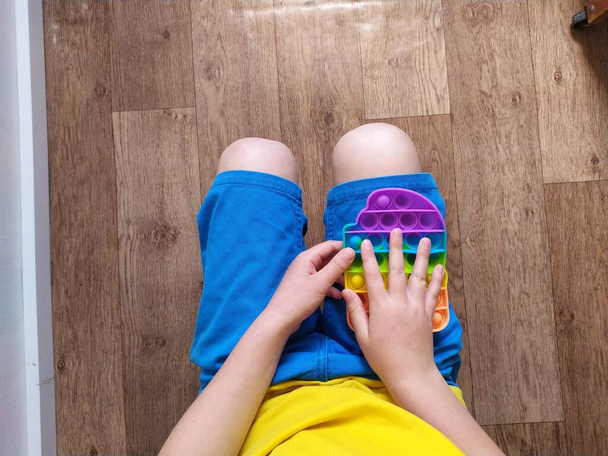 Chłopiec w niebieskich spodenkach bawić się kolorowym poppit fidget zabawki. Antystresowa zabawka sensoryczna dla dzieci i dorosłych - Zdjęcie, obraz