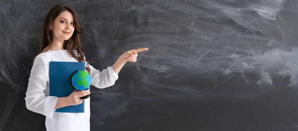 Young teacher or student millennial girl standing with a globe and a notebook in front of a black chalk board and pointing her finger at a blank space for text. Banner. University education concept. - Foto, Imagem