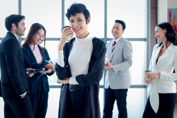 Умная и элегантная улыбающаяся деловая женщина, женщина-босс с черными короткими волосами конфиденциально стоя и держа чашку кофе бумагу на рабочем месте оконного офиса, четыре бизнесмена, как размытый фон. - Фото, изображение