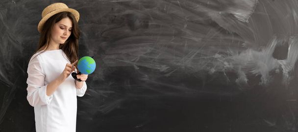 Caucasian girl in a summer hat with a globe standing near an empty chalk board. Summer vacation planning concept. Copy space. banner. - Photo, Image