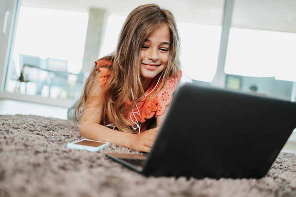 Beautiful girl lies on the floor of the living room of her house and uses a laptop for learning and communication. She is happy and smiled. - Photo, image